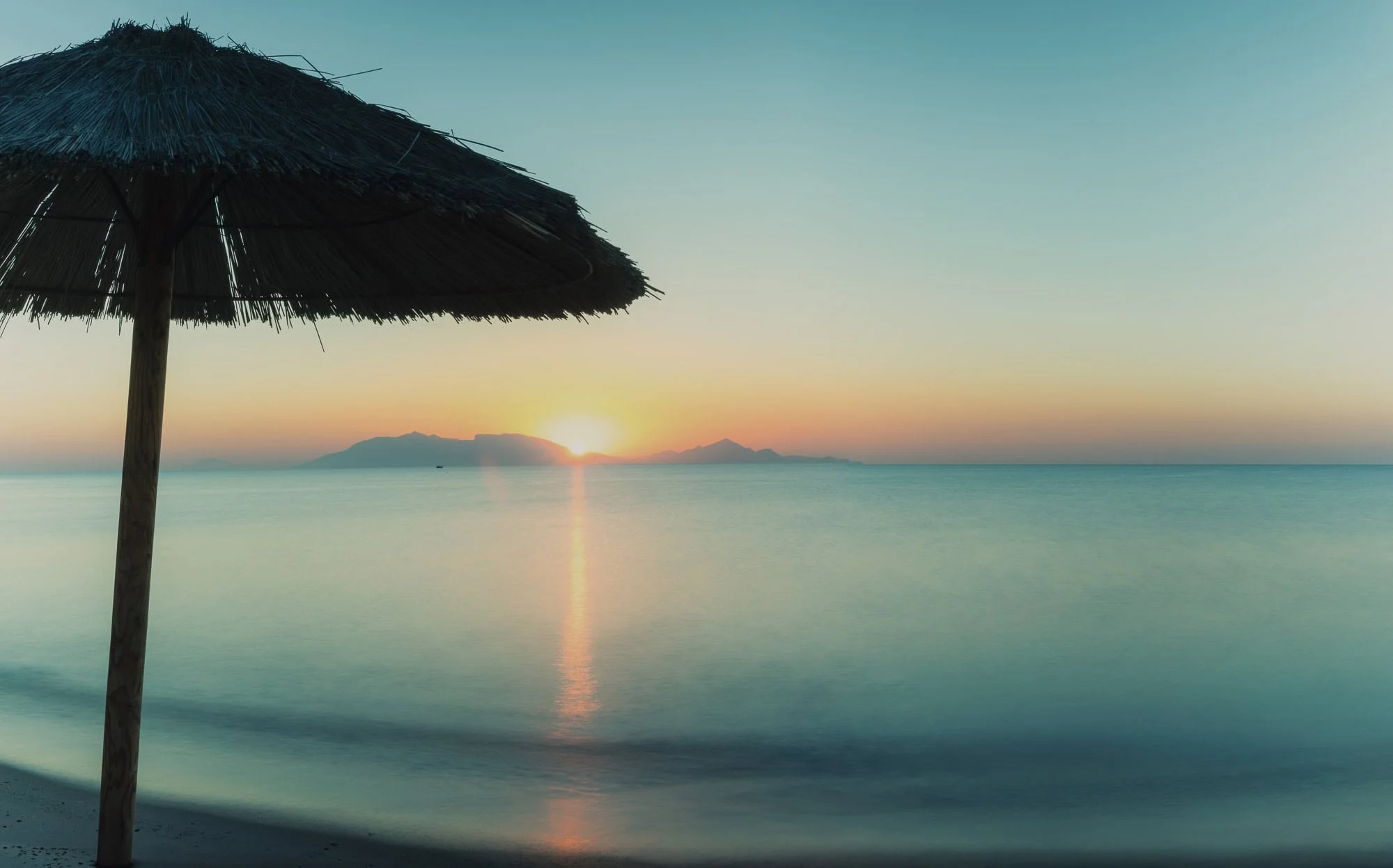 "Umbrella at the beach of Kardamena, Kos, the sun rising over the rugged silhouette of the Turkish mountains, viewed across the sea from the Greek Dodecanese islands. Fine art photography available as a 100% cotton canvas or Limited Edition Giclée print, featuring a 1.5-inch (3.8 cm) solid Latvian pine frame."
